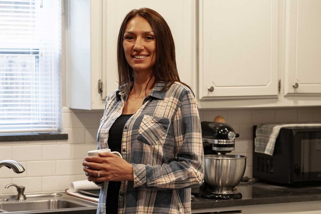 A woman stands in her kitchen smiling, holding a coffee cup. Connection to safe housing is possible with Dragonfly Human Trafficking Victim Services